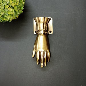 May include: A gold-toned metal door handle shaped like a hand with a decorative band around the wrist.