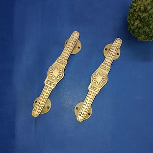 Puede incluir: Dos tiradores de puerta de latón dorado con diseño floral y detalles decorativos. Los tiradores son de color dorado y están sobre un fondo azul. Los tiradores son probablemente para un armario o una puerta.