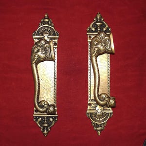 May include: A pair of antique brass door handles featuring an elephant head motif. The rectangular handles have a decorative border and a curved handle resembling an elephant's trunk. The background is a deep red colour.