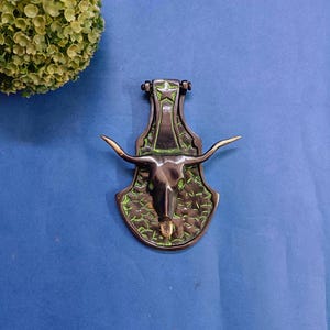 May include: A bronze-colored door knocker featuring a longhorn skull design. The knocker has a star at the top and green accents. The horns are a lighter bronze color. The knocker is mounted on a blue surface.