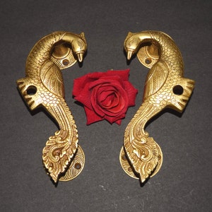 May include: Two gold-toned metal door handles shaped like peacocks with intricate details. The handles are facing each other and are set against a black background.