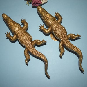 Könnte beinhalten: Zwei goldfarbene Alligator-Türklopfer. Die Alligatoren sind mit strukturierten Schuppen verziert und haben einen offenen Mund.