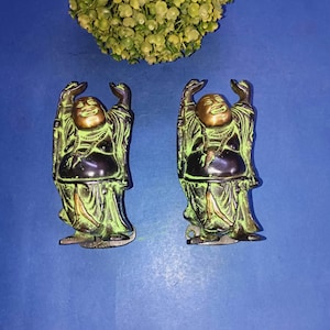 May include: Two bronze-colored laughing Buddha statues with raised arms and a gold-colored head. The statues have a green patina and are set against a blue background. A cluster of green flowers is in the background.