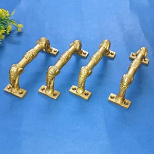 Puede incluir: Cuatro manijas de puerta de latón con diseño de cabeza de elefante. Las manijas doradas tienen intrincadas tallas. Están montadas sobre placas rectangulares y dispuestas sobre una superficie azul.