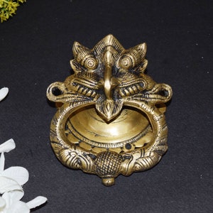 May include: A gold-toned brass door knocker shaped like a mythical creature's head with intricate details. The knocker has a ring for pulling and a detailed face with large eyes.