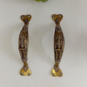 May include: A pair of ornate brass door handles with intricate designs featuring a seated figure. The handles have a heart-shaped top and a curved, elongated body.