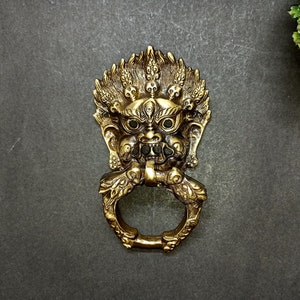 Puede incluir: Un llamador de puerta dorado en forma de cara de criatura mítica con un anillo para el asa.