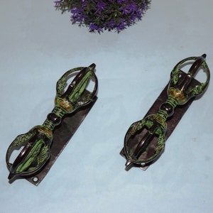 May include: Two antique bronze vajra scepters with green and gold accents, resting on dark rectangular bases. The scepters have intricate, ornate designs and are set against a light blue background, with purple flowers in the top corner.
