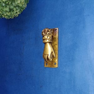 May include: A gold-toned door knocker shaped like a hand with intricate details. The knocker is mounted on a rectangular brass plate.