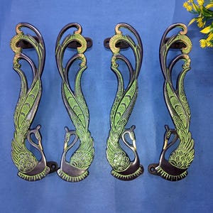 Może przedstawiać: Cztery dekoracyjne klamki do drzwi z motywem pawia. Klamki są czarne z zielonymi i złotymi akcentami. Każda klamka ma motyw pawia i łabędzia, dodając eleganckiego akcentu do każdych drzwi.