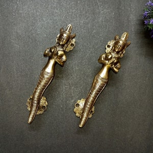 Puede incluir: Dos tiradores de puerta dorados en forma de mujeres con tocados ornamentados y cuerpos detallados. Los tiradores están sobre un fondo gris.