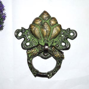 May include: An ornate bronze door knocker with a detailed design. The knocker features a central floral motif with a green patina, flanked by stylized dragon heads. The ring handle is also bronze.