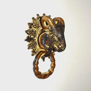 Puede incluir: Un aldaba de puerta en tono bronce que representa una cabeza de carnero con tallas detalladas. La cabeza del carnero tiene cuernos curvados y una superficie texturizada. La aldaba incluye un anillo decorativo y un diseño de hojas detallado.