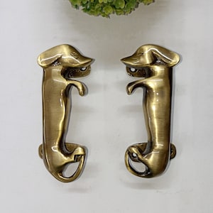 Può includere: Un paio di maniglie per porte in ottone a forma di bassotto. Le maniglie sono in posizione eretta con le gambe piegate e le code arrotolate.