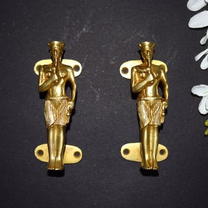 Puede incluir: Dos tiradores de puerta dorados en forma de figuras de pie. Cada tirador presenta un diseño detallado de una persona con un tocado y sosteniendo un bastón.