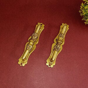 May include: Two ornate brass drawer pulls with a decorative floral design. The pulls are a light gold color and have a slightly aged finish.