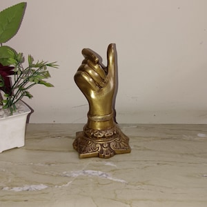 Könnte beinhalten: Eine goldfarbene Metallhandskulptur mit einem erhobenen Zeigefinger, der nach oben zeigt. Die Hand befindet sich auf einem dekorativen Sockel mit filigranen Details.