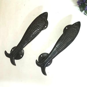 Op de afbeelding: Twee zwarte, visvormige deurgrepen. De metalen handgrepen hebben een gedetailleerd schubbenpatroon. Elk heeft een staartvin en een ronde basis met schroefgaten. Decoratieve deurbeslag.
