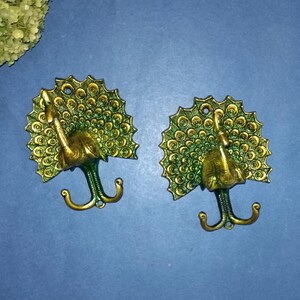 May include: Two brass peacock-shaped wall hooks. The hooks have a detailed peacock design with a fan-shaped tail and a curved hook. The hooks are in a brass color with green accents, set against a blue background.