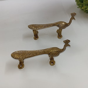 Könnte beinhalten: Zwei goldene Türgriffe in Form eines Pfaus mit aufwendigen Details. Die Griffe sind aus Metall und haben eine polierte Oberfläche.