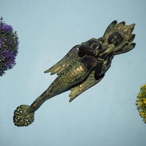May include: A bronze mermaid door handle with detailed craftsmanship. The mermaid features wings and a scaled tail, with an ornate handle end. The handle is set against a light blue backdrop, with purple and yellow plants.
