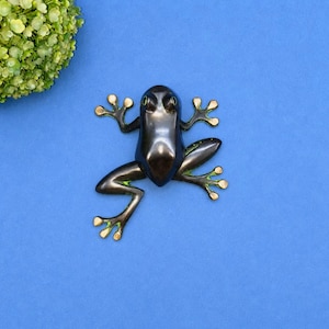 May include: A bronze frog figurine with green eyes and accents, posed on a blue background. The frog's feet and the edges of its legs are painted with a light green color. The frog is positioned in the center of the image.