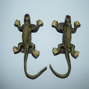 May include: Two black and green lizard-shaped hooks with a gold finish. The hooks are mounted on a wall and have a curved tail that forms a hook.