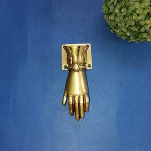 Peut inclure: Un heurtoir de porte en laiton en forme de main. Le heurtoir est monté sur une plaque rectangulaire avec un accent ovale. La main est en position basse avec des doigts détaillés. La finition est de couleur or brillant.