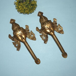 May include: Two gold-toned metal door handles with a decorative design featuring a face with a third eye. The handles are shaped like a stylised human figure with a long, curved spout.