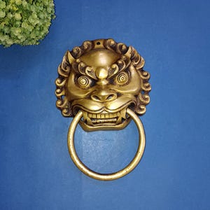 May include: A gold lion's head door knocker with a circular ring. The knocker is detailed with a fierce expression and intricate carvings.