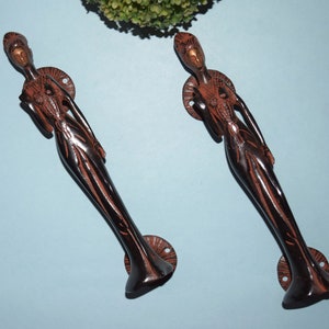 May include: Two ornate, dark brown, metal door handles shaped like a standing woman. The handles have a gold finish on the face and are mounted on a round base.