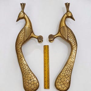 Puede incluir: Dos manijas de puerta doradas con forma de pavo real. Las manijas son de metal y presentan detalles intrincados, incluido un cuerpo texturizado y una cabeza detallada. Se coloca una regla amarilla entre las manijas.