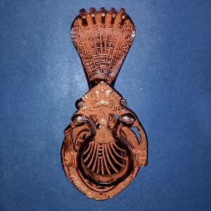 May include: A decorative wall hook made of metal with a brown finish. The hook is shaped like a cobra with its head raised and a ring in its mouth. The cobra is adorned with intricate details and a gold-colored crown.