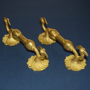 May include: Two ornate, gold-colored door handles. Each handle features a bird-like design with a curved body and a decorative base. The handles are made of metal and have a vintage aesthetic, suitable for antique or classic decor.