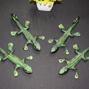 May include: Four green metal lizard figurines with textured details and a bronze finish. The lizards are arranged on a dark surface, with a small white pot of yellow flowers in the background. Decorative reptile figurines.