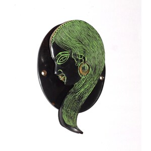 Puede incluir: Adorno de pared negro con el perfil de una mujer con largo cabello verde. La cara y el cabello están grabados con líneas verdes sobre un fondo negro. La pieza tiene forma circular con tres pequeños agujeros.