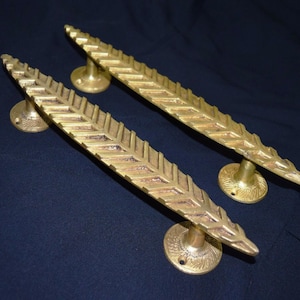 May include: Two brass-coloured door handles with a leaf design. The handles are long and slender, with a series of angled lines creating a feather-like pattern. Each handle has a circular base for mounting. The handles are on a dark blue background.