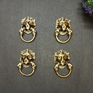 May include: Four gold lion head door knobs with a ring pull. The lion heads are detailed with a mane and a closed mouth.