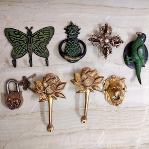 May include: A collection of seven decorative metal wall hooks and door knockers including a green butterfly, a pineapple door knocker, a bronze vajra, a green parrot, a padlock with a key, two gold lotus flower hooks, and a gold lion head.