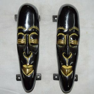 May include: Two black decorative door handles with a carved tribal mask design. The masks have gold-colored accents on the eyes, nose, and mouth. Each handle has a rectangular metal mounting plate.