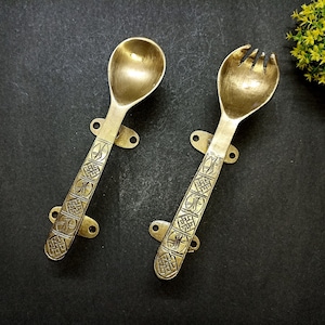 May include: A pair of decorative brass serving utensils, a spoon and a fork, with ornate, engraved handles. Each utensil has a mounting bracket with two holes for hanging. The background is a dark gray surface.