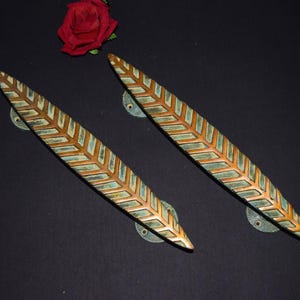 May include: A pair of bronze-effect door handles, each with a leaf design and a geometric pattern in green and gold. The handles are set against a dark background, with a red rose in the upper left corner.