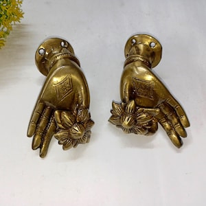 Puede incluir: Dos aldabas antiguas de latón con forma de mano. Cada aldaba presenta una mano detallada con una flor en la muñeca y un patrón de diamante decorativo en la palma. Las aldabas están montadas sobre una base redonda con dos agujeros.