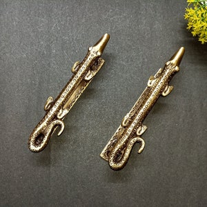 Puede incluir: Dos tiradores de puerta de metal dorado con forma de cocodrilos. Los tiradores están detallados con escamas texturizadas y tienen una forma ligeramente curvada.