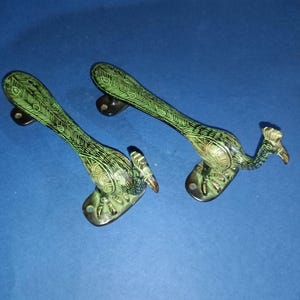May include: Two decorative peacock-shaped door handles. The handles are made of metal with a green and bronze finish. The peacock design includes detailed feather patterns and a small beak. The handles are mounted on a dark base.