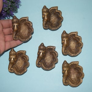 Könnte beinhalten: Sechs antike, goldfarbene dekorative Öllampen, jede mit einer detaillierten Darstellung der hinduistischen Gottheit Ganesha. Die Lampen sind auf einer hellblauen Oberfläche angeordnet, mit einer Hand, die eine der Lampen in der Nähe einer lila Blumengestecke hält.