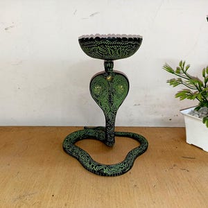 May include: A decorative, green and black cobra-shaped candle holder. The candle holder has a bowl-shaped top with a detailed leaf pattern. The cobra's head is upright, and the body forms a loop at the base. The candle holder is made of metal.