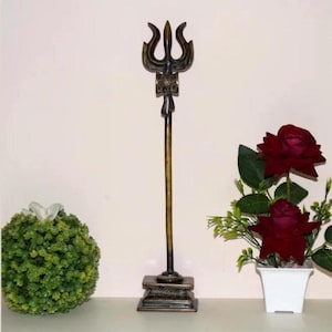 May include: A bronze-colored trident statue with a square base, displayed on a white surface. The trident is flanked by a green topiary ball and a small white pot with red roses. The statue is a decorative item.