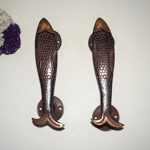 以下が含まれることがあります： テクスチャ加工された表面を持つ、黒と金の魚の形をしたドアノブ2つ。ノブは金属製で、装飾的なデザインが施されています。