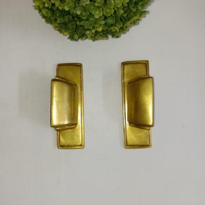 Puede incluir: Dos tiradores de puerta de armario de color dorado con forma rectangular y una sección central ligeramente curvada en relieve.